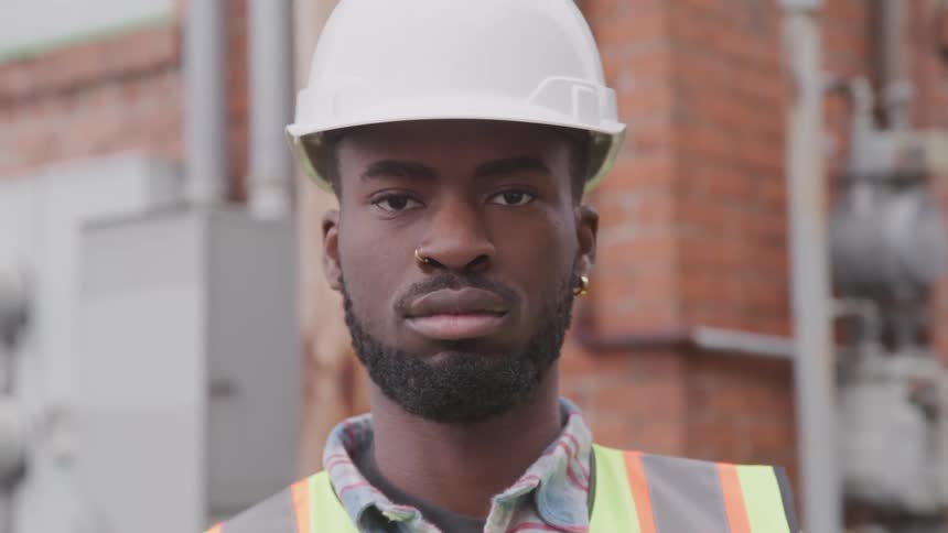 A young black man in a vest and hard hat in front of a building