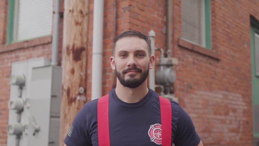A firefighter wearing suspenders and a t shirt standing in front of a building