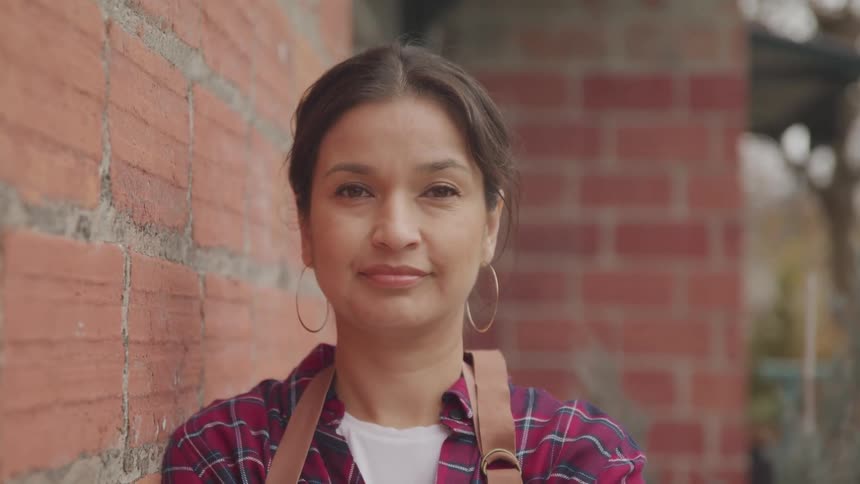 A middle aged hispanic woman smiles into the camera while leaning against the facade of a brick building