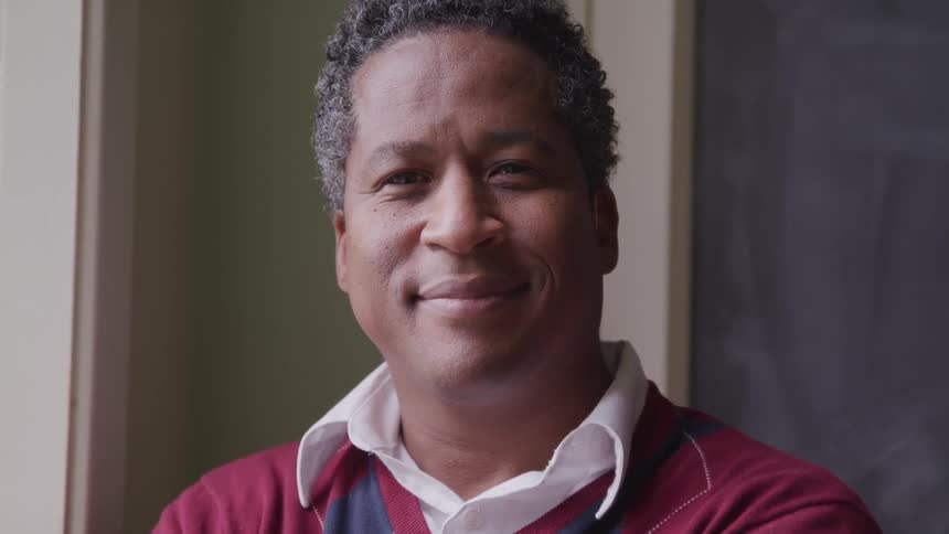 Middle aged man with a shirt and a red sweater smiles slightly and looks at the camera