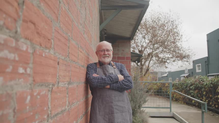 Elderly man in apron leans on brick wall with arms crossed and smiling