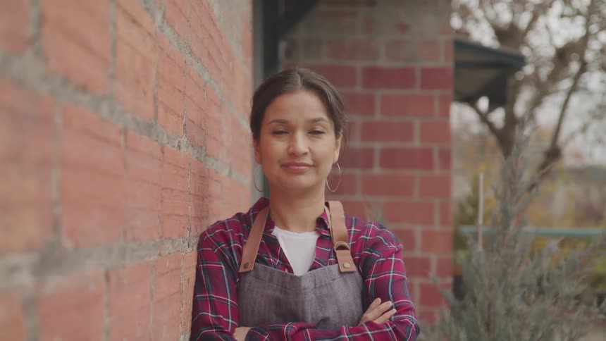 A hispanic woman with her arms crossed and a shoulder resting against a building looks into the camera with a soft smile