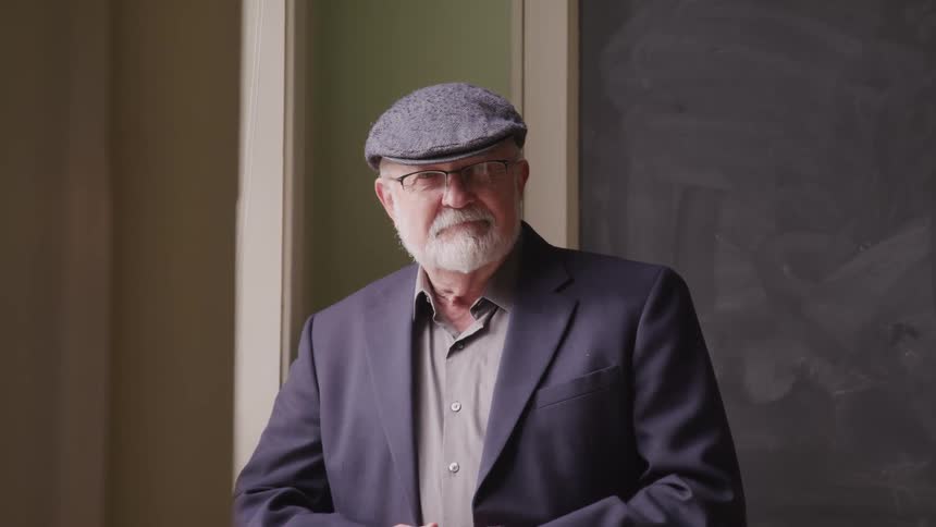 Elderly man with a flat cap a grey beard and jacket stands by window and faces the camera with eye contact