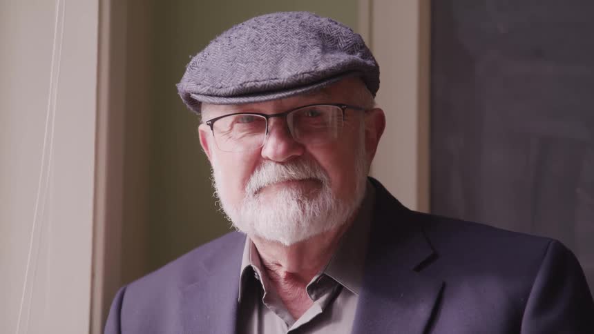 An elderly white man with white facial hair and a gray hat and coat is standing near a window looking ahead