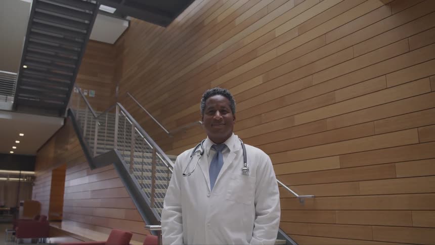 Dark skinned man with a white lab coat and stethoscope looks at the camera smilingly in the hospital