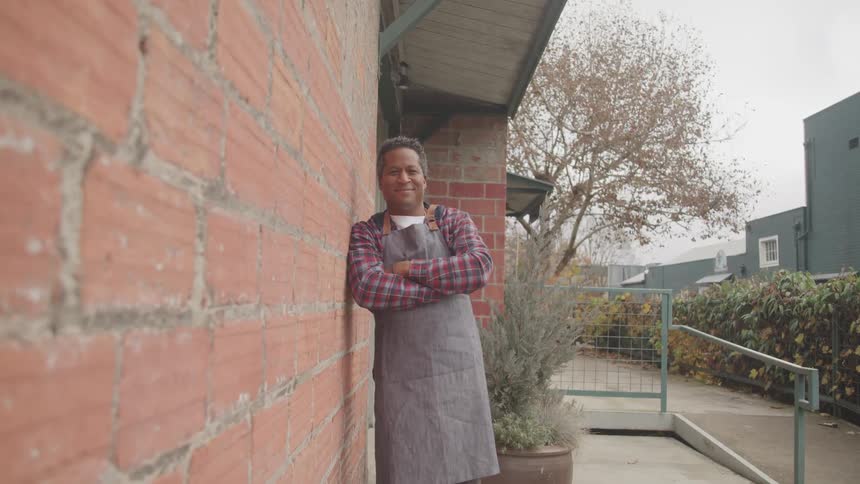 A man wearing an apron stands against the outside of a brick building