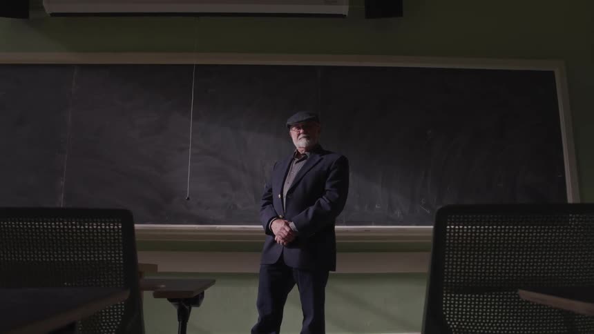 A man with a hat and jacket standing in front of a classroom with a blackboard in the background