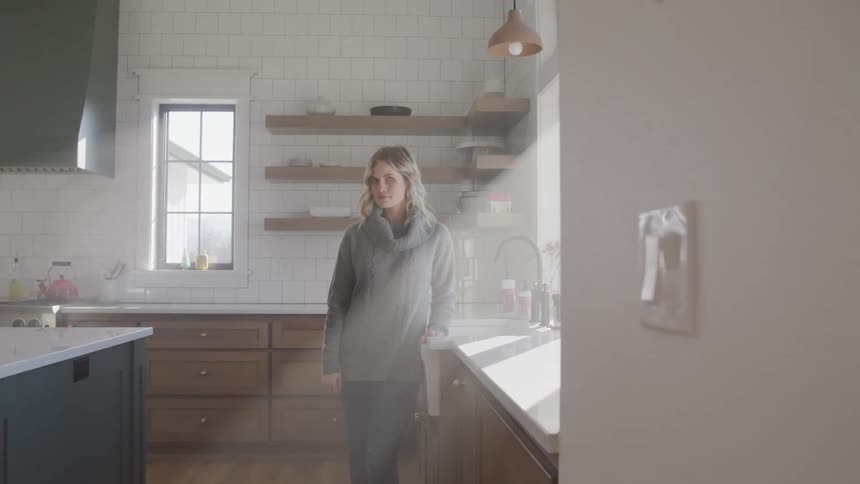 Blonde woman with a grey sweater stands by a counter in the kitchen