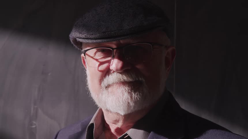 Older white bearded man wearing golf cap and eyeglasses stands in front of gray wall