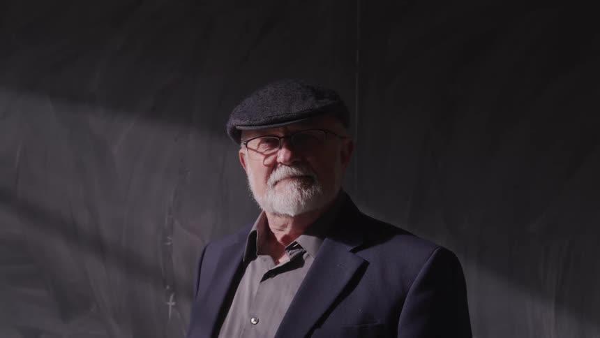 Elderly man with a flat cap a beard and glasses faces the camera with eye contact in front of a blackboard