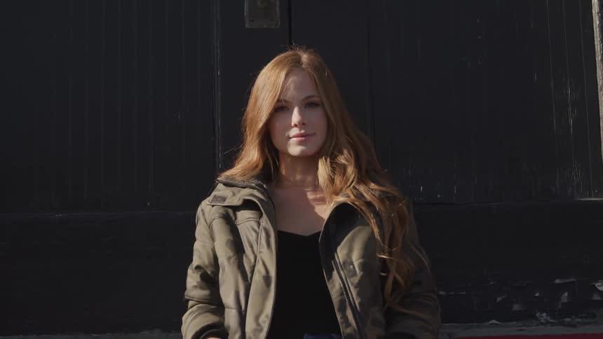 Woman with red long hair a green coat stands in front of a black exterior door of a building and turns her head to the camera