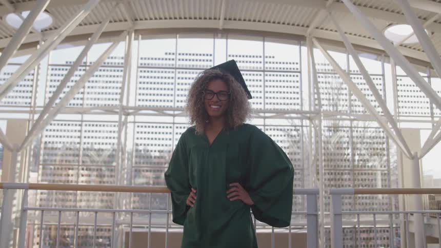 A woman stands on an interior balcony wearing a graduation cap and gown