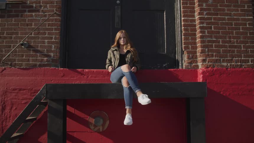 A woman sitting on a wooden platform at the top of a small staircase outside of a building turns her head to look into the camera