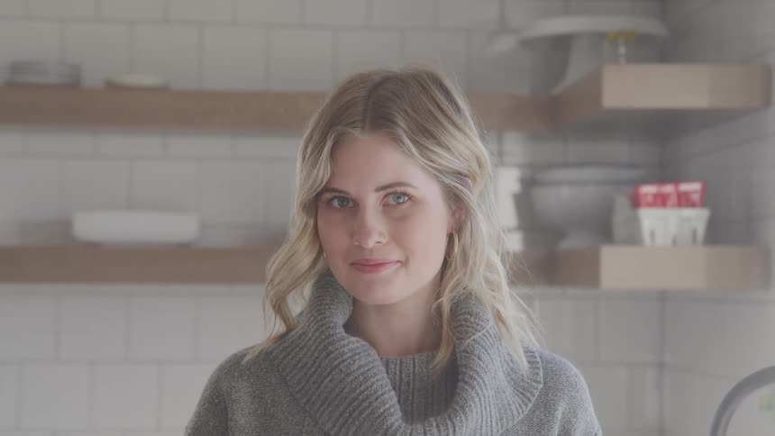 Blonde woman with a grey sweater faces the camera with eye contact in the kitchen