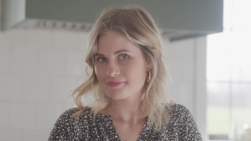 A young woman takes her gaze to look into the camera while sitting a kitchen