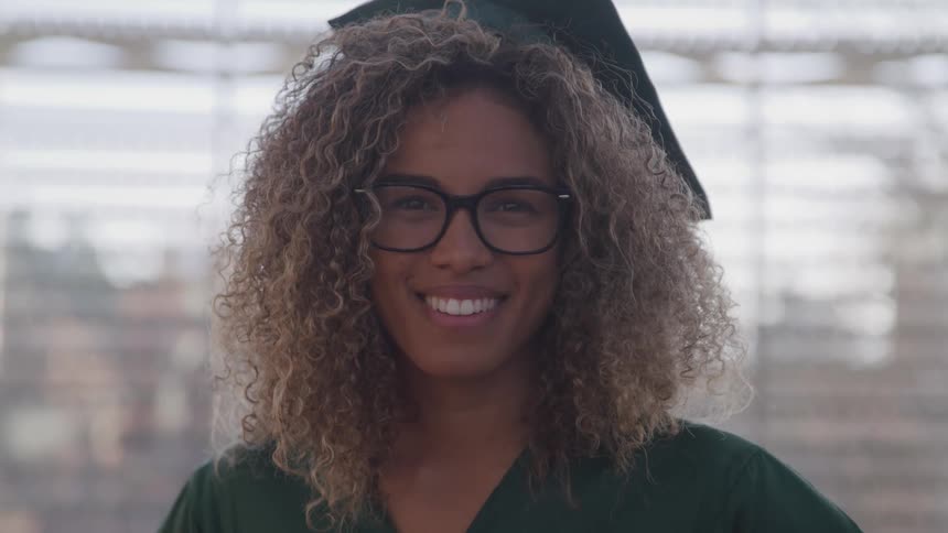 A black with brown curly hair is woman wearing a cap and gown and a big smile on her face