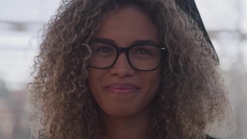 A young black woman with glasses smiles sweetly as she looks into the camera