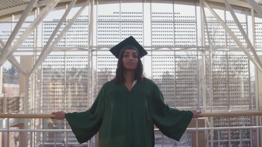 A woman stands on an interior balcony wearing a graduate cap and gown