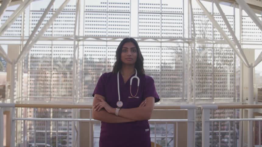 An indian woman wearing purple scrubs is standing with her arms cross posing in front of a railing in a large building
