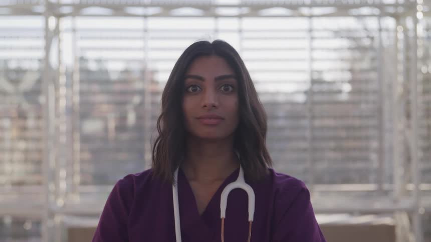 Dark skinned female doctor with purple medical uniform and stethoscope faces the camera with eye contact