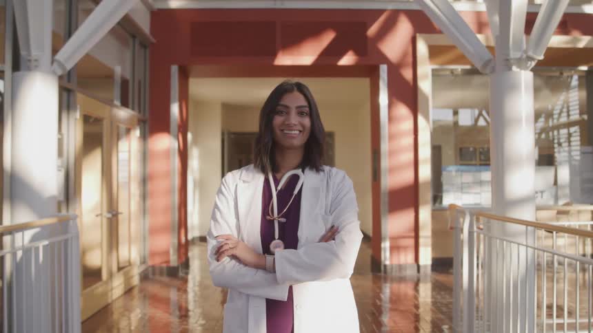 Brunette female doctor with a stethoscope and a white lab coat stands arm crossed and looks at the camera smilingly