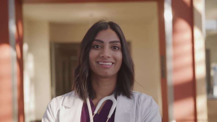 Dark skinned female doctor with a white lab coat a purple medical uniform and stethoscope faces the camera with eye contact