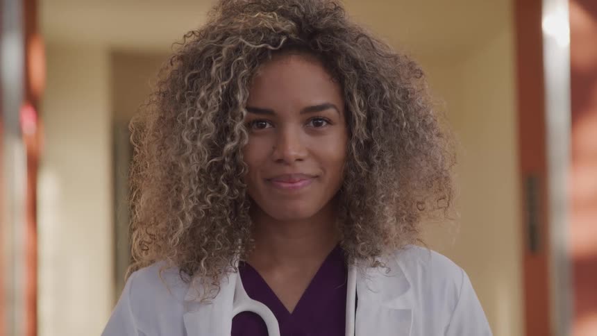 Brunette female doctor with curly hair a stethoscope and a white lab faces the camera with eye contact smilingly