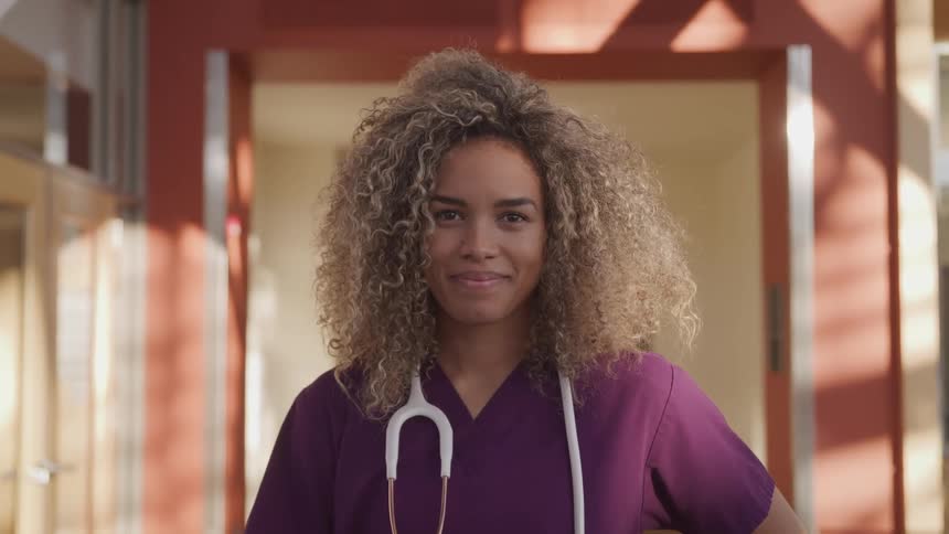 A nurse stands inside a building with a stethoscope around her neck and smiles