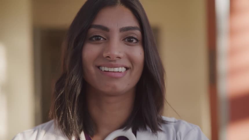 Brunette female doctor with a stethoscope and a white lab coat turns her head and smiles at the camera