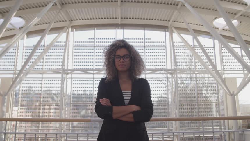 A young black woman with her arms crossed turns her head to look into the camera