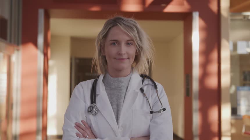 A female doctor smiles into the camera while standing in a corridor with her arms crossed