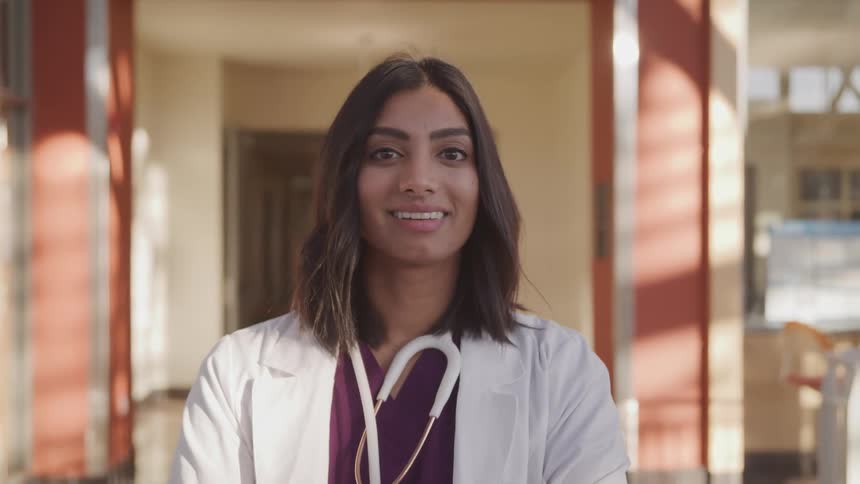 Dark skinned female doctor with a white lab coat purple medical uniform and stethoscope faces the camera smilingly with eye contact