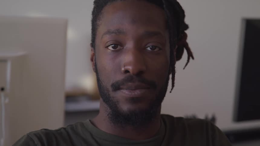 Black man with braid hair a beard and a nose ring hoop faces the camera with eye contact