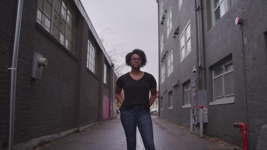 A woman stands with her hands in her back pockets as she poses in the alley between two buildings in a city