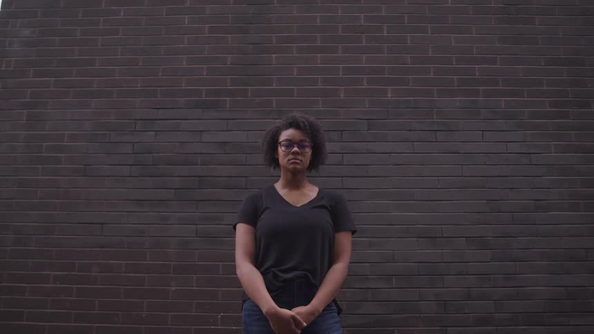 Black woman with curly hair and glasses stands and stares at the camera in front of a brick wall