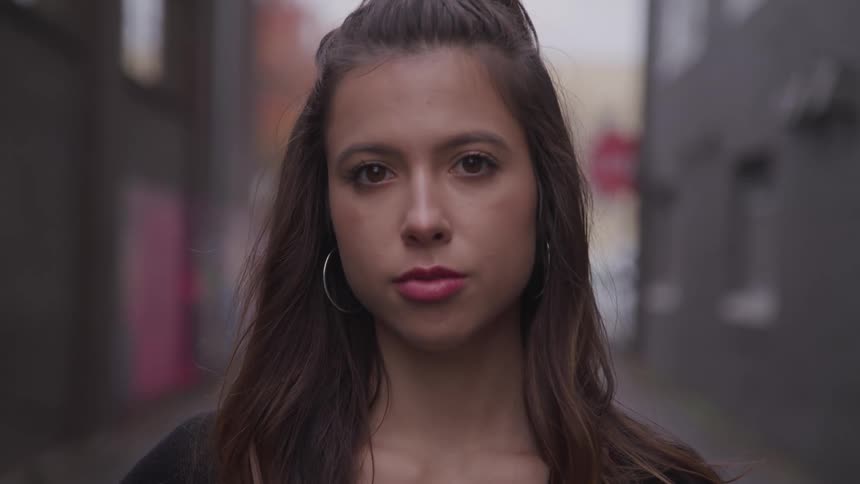 Brunette woman with brown hair and hoop earrings faces the camera with eye contact in the alley