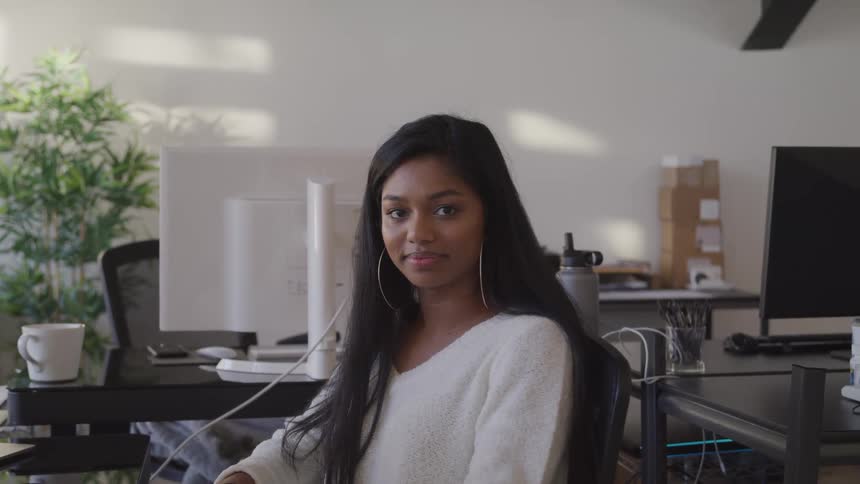 Black woman with a white sweater and hoop earrings sits in a chair and looks at the camera in an office