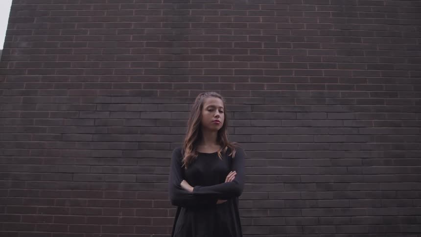 Woman with brown hair and a black shirt stands arms crossed in front of a brick wall