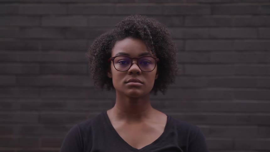 A black girl with short black curly hair and glasses wearing a black shirt is standing in front of a black brick wall