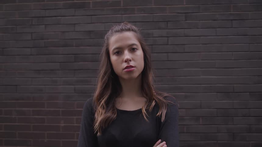 A young woman stands with her arms crossed as she poses in front of a brown brick wall