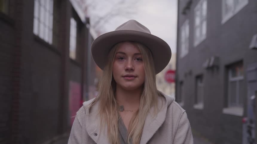 Blonde woman with a hat and overcoat stands in a narrow street on a cloudy day