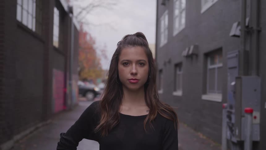 A woman stands in an alleyway between two buildings and stares into the camera on a cloudy autumn day