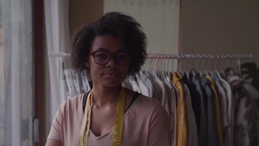 A young black woman with short curly black hair and glasses is standing with a tape measure around her neck next to a clothes rack