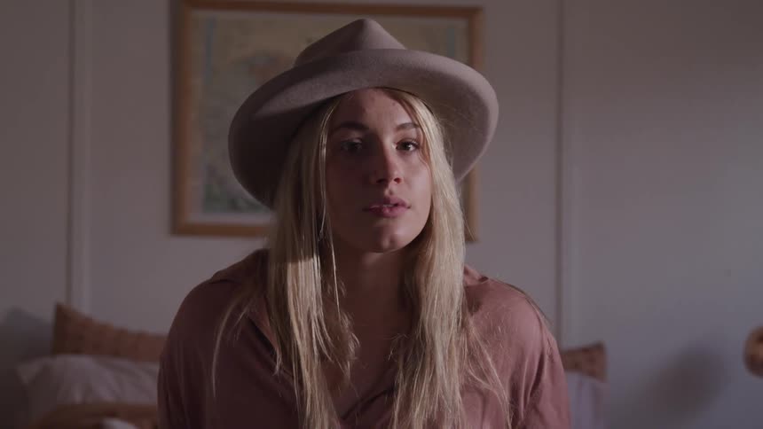 Blonde woman wearing hat sits on bed