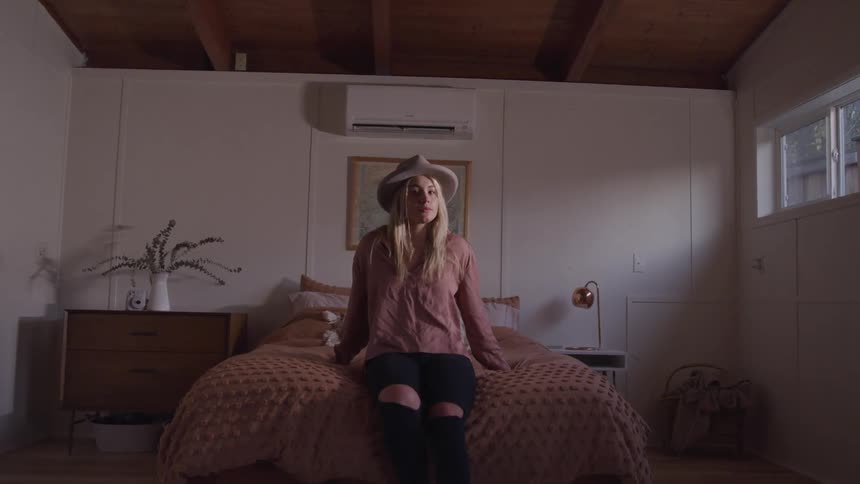 A young woman sitting on a bed in a bedroom turns her head to look into the camera