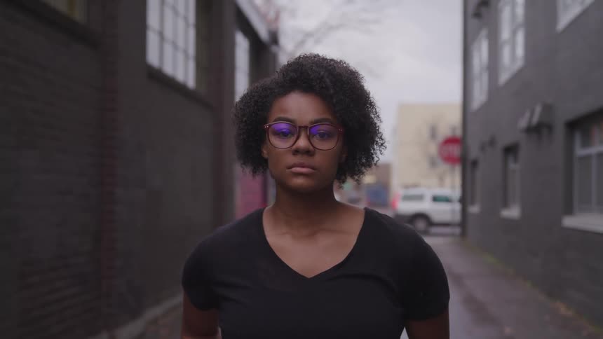 A woman with glasses standing and staring in an alley with buildings in the background