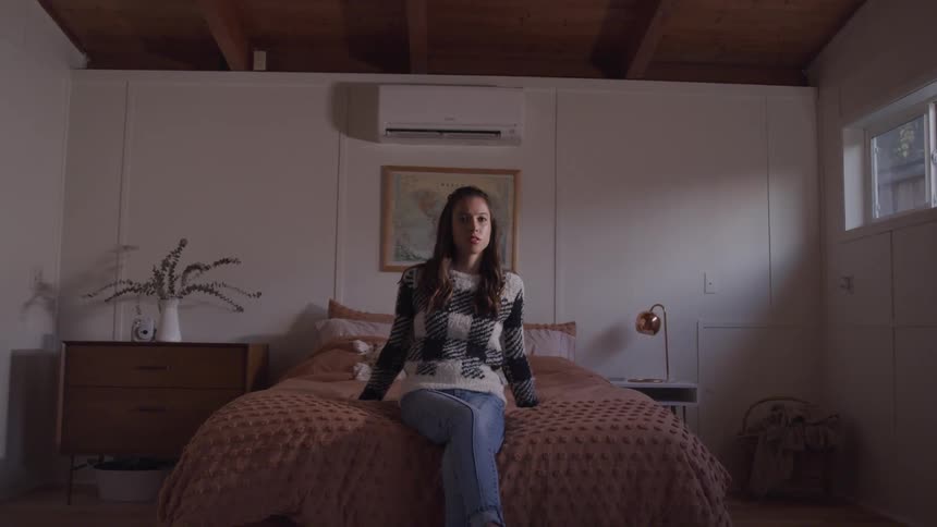 Woman with a sweater and jeans sits on bed and looks at the camera in bedroom