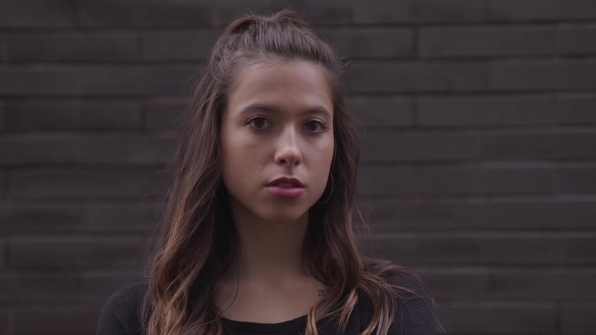 Young woman with long hair standing in front of a brick wall turns to look at the camera