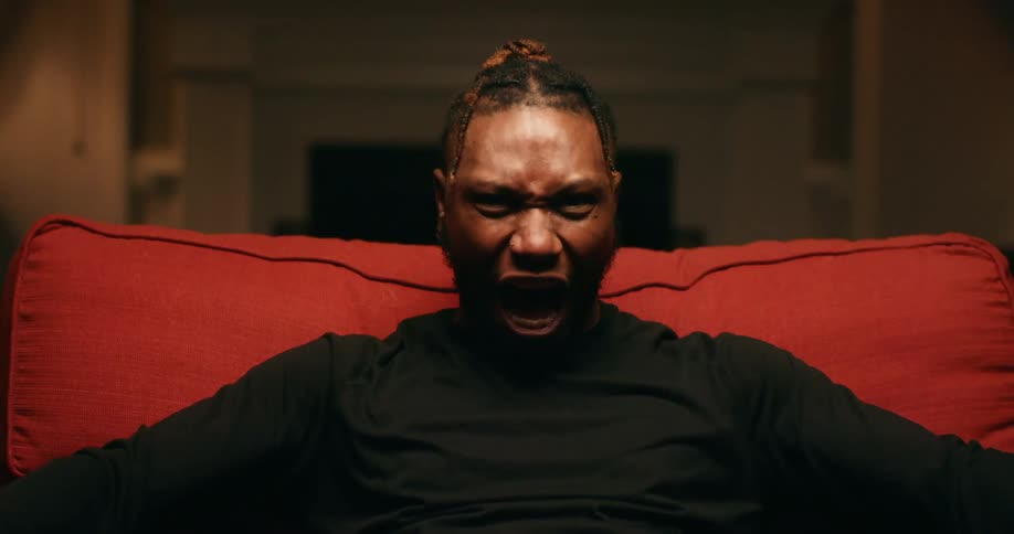 A black man with braids wearing a black long sleeve shirt is sitting on a red chair yelling angrily