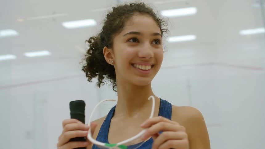 Woman stands next to clear wall smiling as she holds a racket and glasses